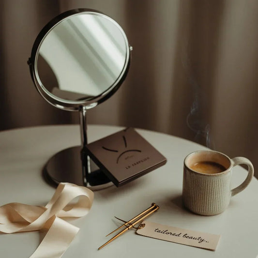 Flatlay of a premium vanity setup with a brown lash tray, gold tweezers, ribbon, ceramic coffee mug, and mirror on a neutral table – styled to reflect tailored beauty and elegance for a lash extension guide.