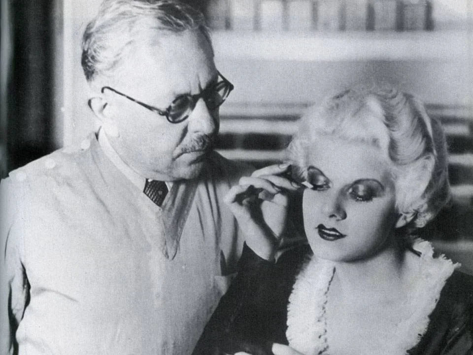 Black-and-white photo of a 1930s Hollywood makeup artist gently attaching false lashes to an actress. The scene reflects the early cinema era that popularised modern eyelash enhancement techniques.