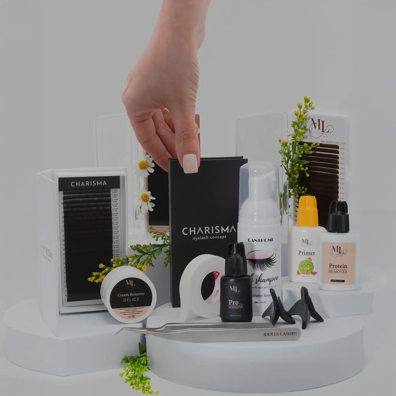 Flat lay of eyelash extension accessories including tapes, glue, primer, remover, tweezers, shampoo, and lash trays, with a hand reaching for CHARISMA box.